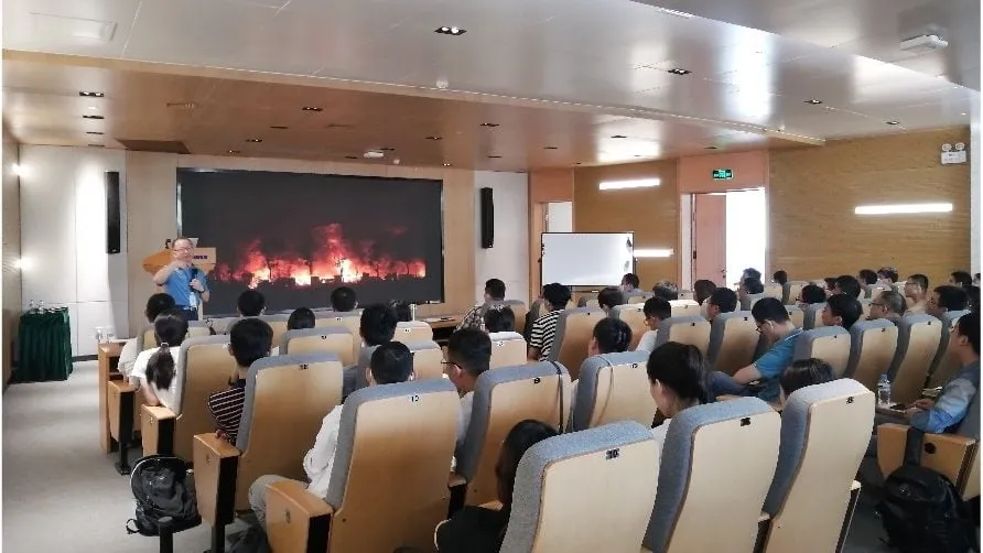 Presenter speaking to an auditorium audience, with a fire video on the screen